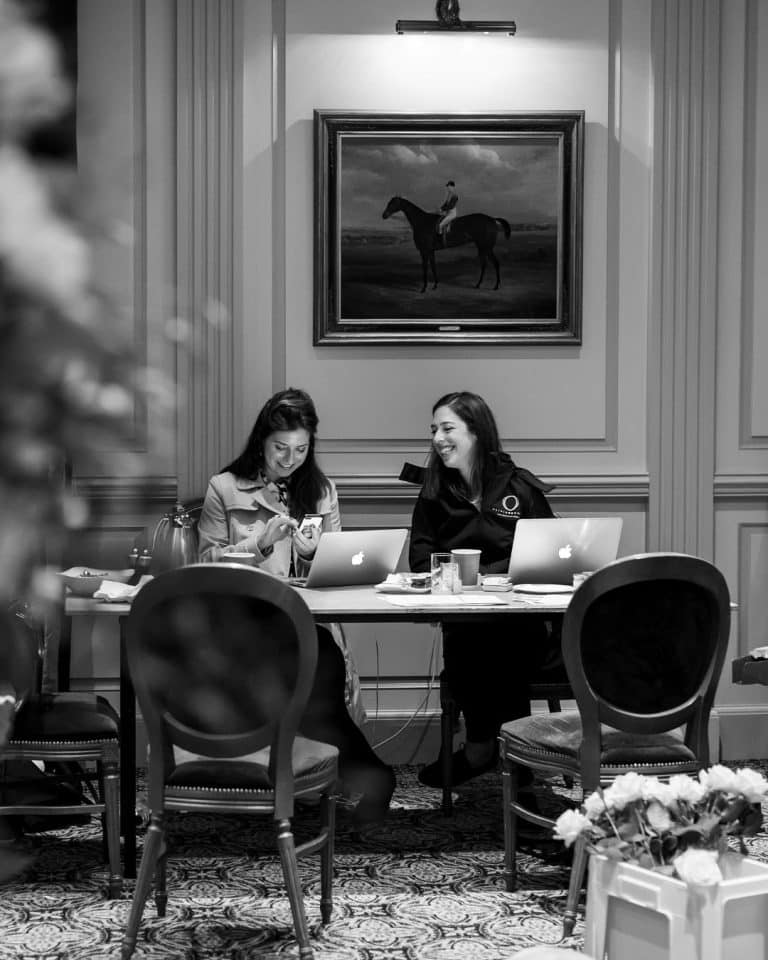 Two women work at a table with laptops, smiling as they chat in a refined room with a large horse-painting on the wall behind them.