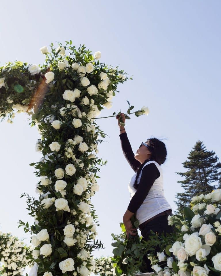 Woman in a white top adjusts white rose flowers on a tall floral arch outdoors under a clear blue sky.