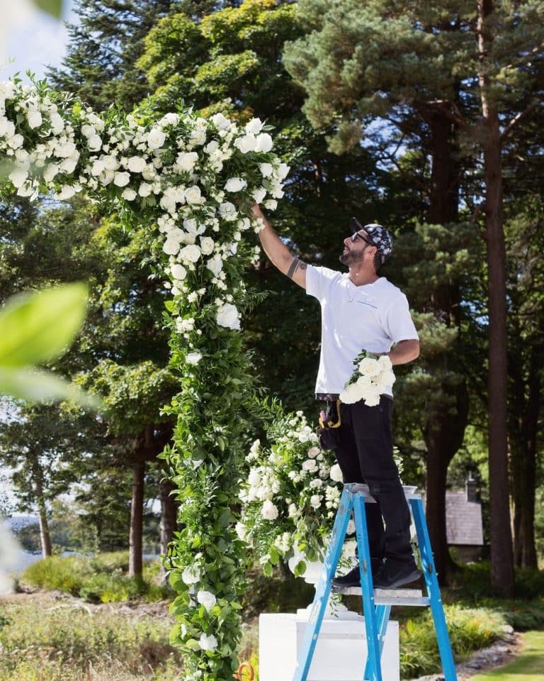 Man on a blue ladder decorating a tall floral arch with white roses and lush greenery outdoors.