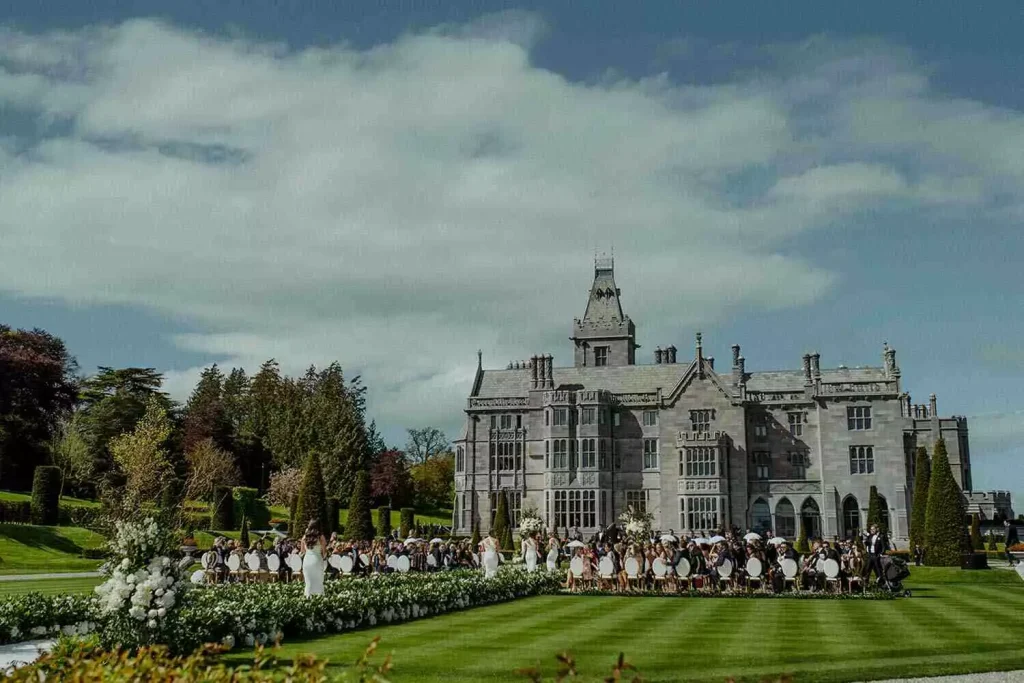 Outdoor wedding ceremony on a manicured lawn in front of a gray stone manor, with rows of chairs and white floral arrangements along the aisle.