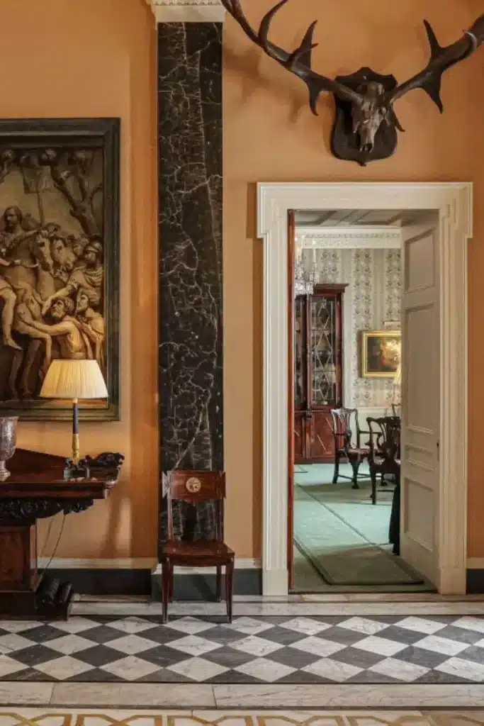 Irish destination wedding Ballyfin Demesne, County Laois, Ireland — foyer with black-and-white checkered tiled flooring, framed painting on the wall, and antler wall decoration above, featuring a wooden side table and wooden chair arranged within the historic entrance hall.
