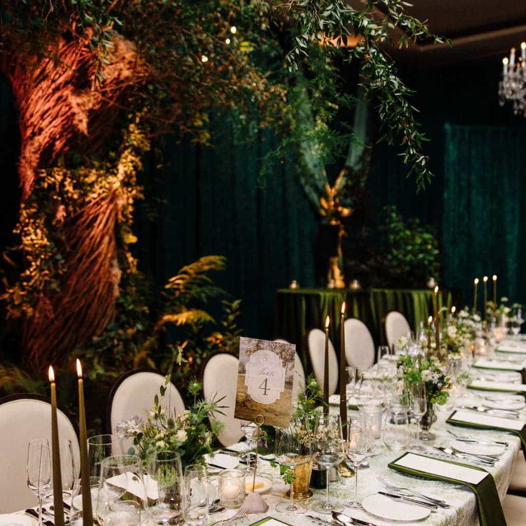 Destination wedding in Ireland at Cashel Palace, County Tipperary — elegant wedding dinner setting with a green and white theme, green napkins topped with printed menus, candlelight, floral arrangements, and table numbers, with a bespoke willow tree installation in the background.