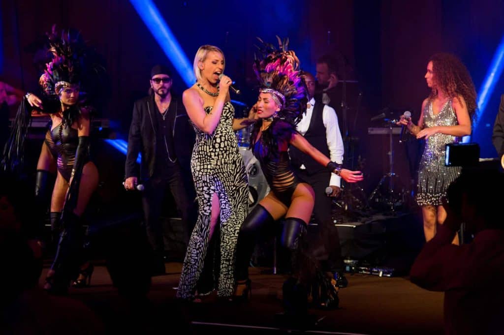A female singer in a black-and-white patterned jumpsuit performs center stage with a microphone while backup dancers in feathered headdresses pose around her under colorful stage lights.