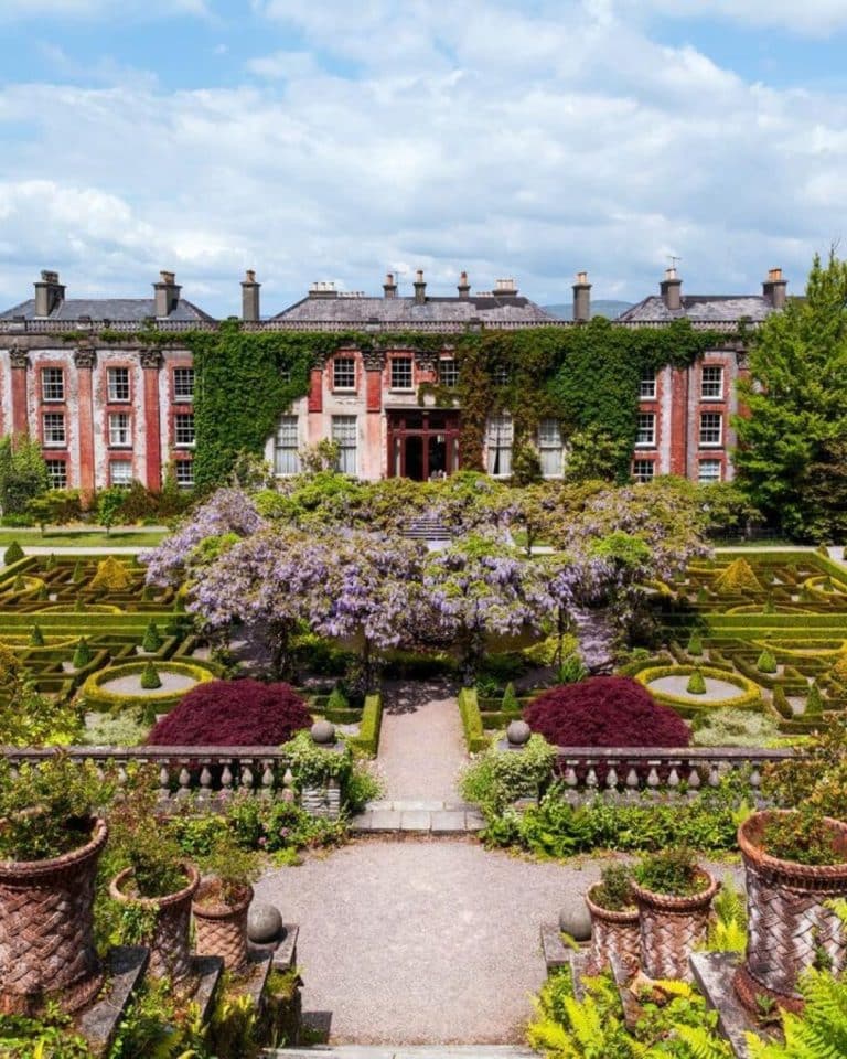 Red-brick manor covered in ivy overlooking a formal, symmetrical parterre garden with trimmed hedges and blooming trees.