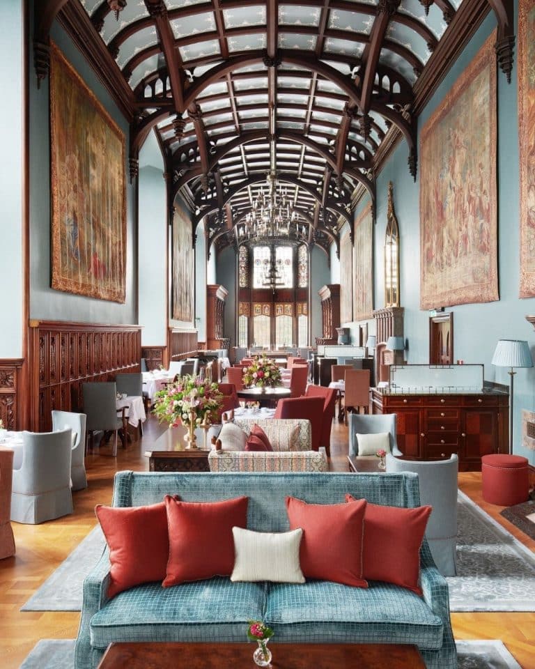 Elegant hotel lounge with teal walls, dark wood arches, and chandeliers, lined with tufted couches and large tapestries.