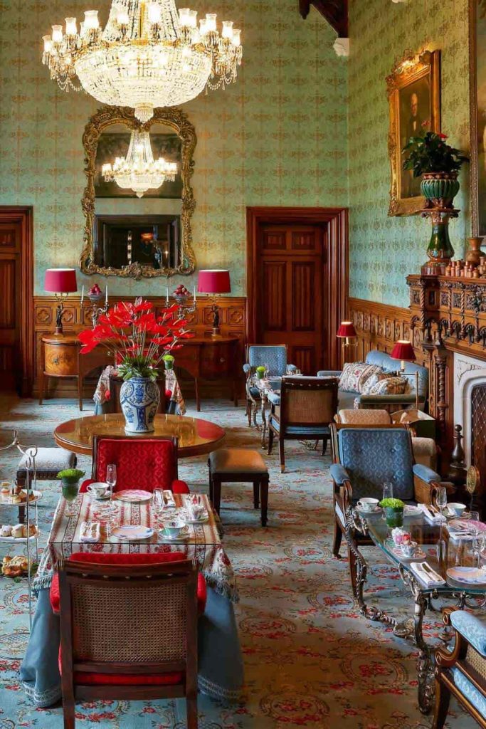Opulent dining/parlor room with a crystal chandelier, ornate mirror, and antique wooden furniture surrounding a table set for tea. A vase of red flowers sits at the center.