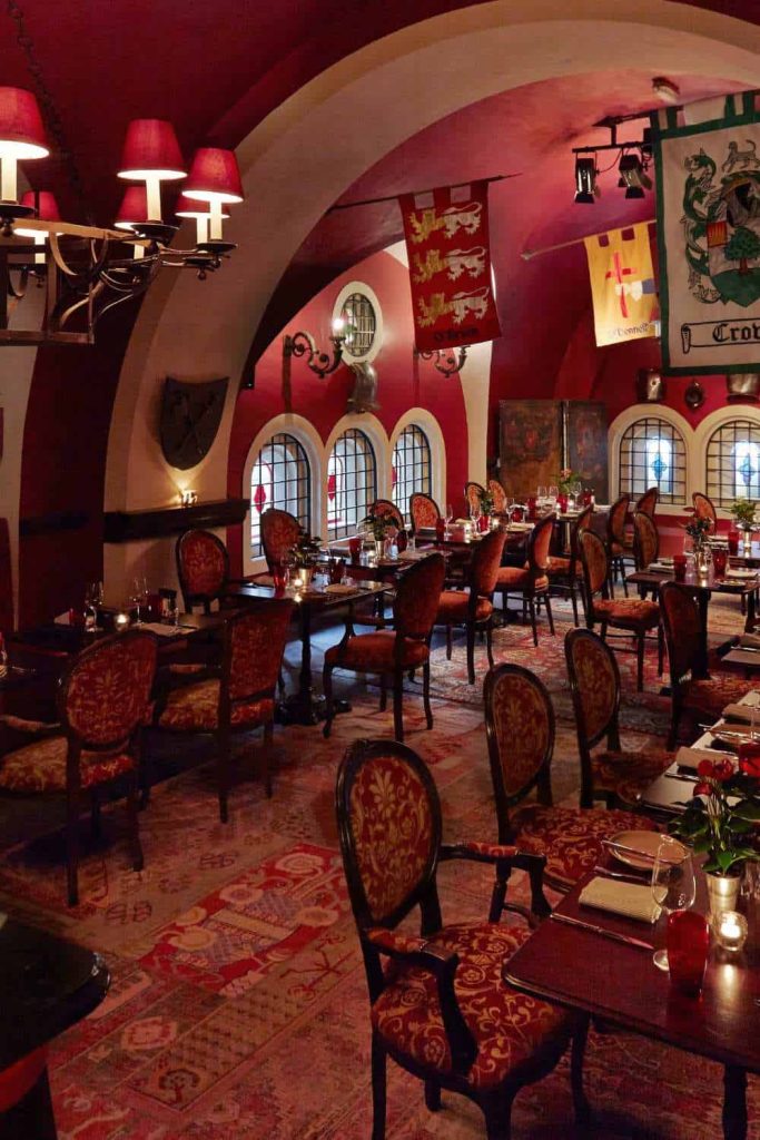 Irish castle wedding at Ashford Castle — The Dungeon dining room with red-themed interiors, set dinner tables, family crest flags on the walls, and arched windows with metal bars.