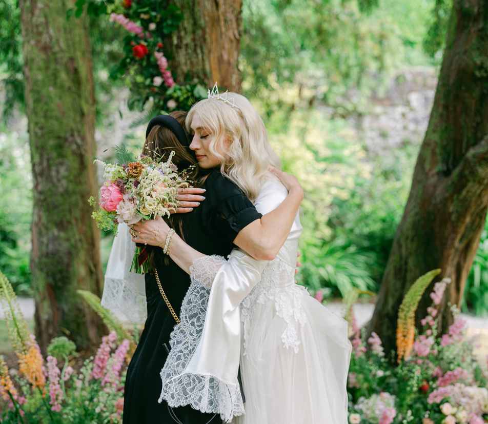 Bride in a white lace dress embraces a guest in a garden setting, bouquet visible between them.