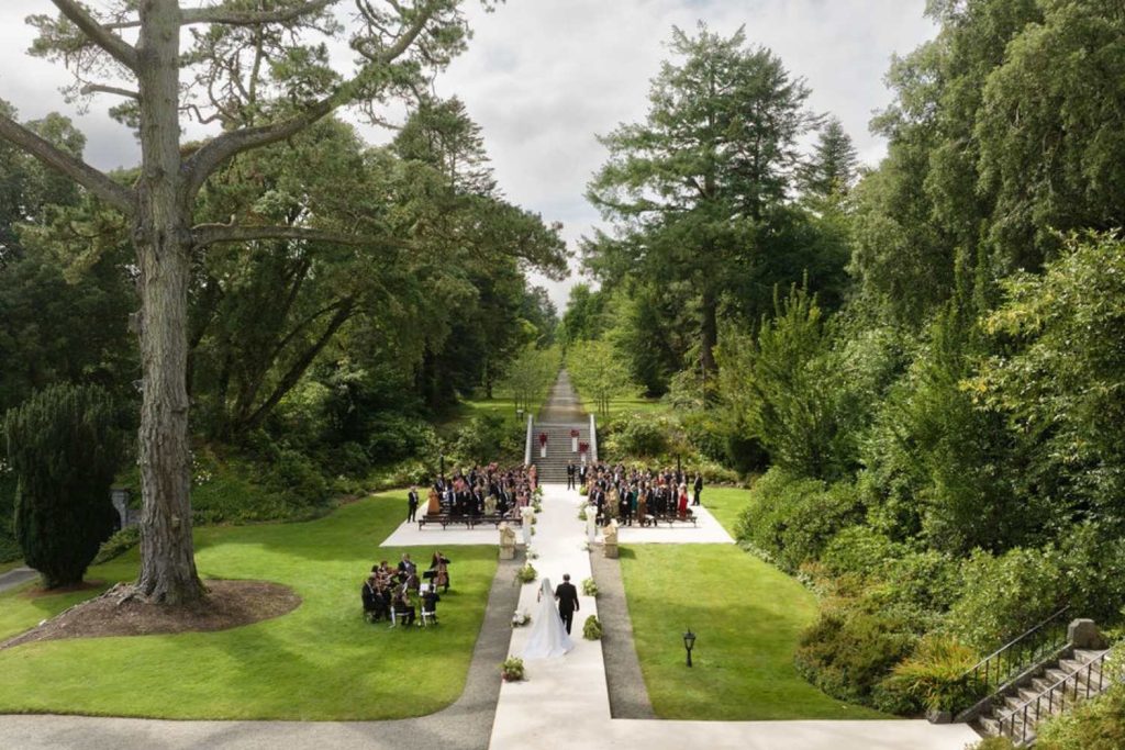 Outdoor garden wedding ceremony: bride and groom walk down the white aisle as guests sit on both sides amid green trees and lawns.