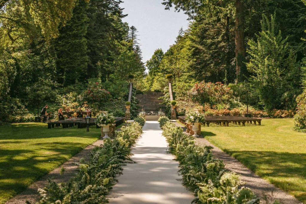 Garden wedding aisle: white runner lined with white flowers leading to stone steps in a sunlit park.