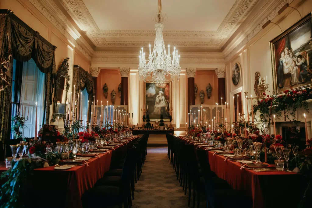 Opulent dining hall set for a banquet with two long candlelit tables and crystal chandeliers across the room.