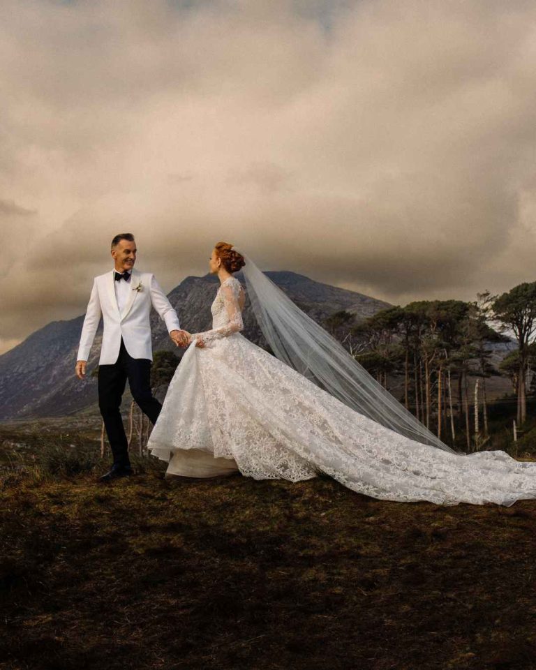 Bride in a lace wedding gown with a long train and veil walks hand in hand with a groom in a white tuxedo against a mountainous landscape under a cloudy sky.