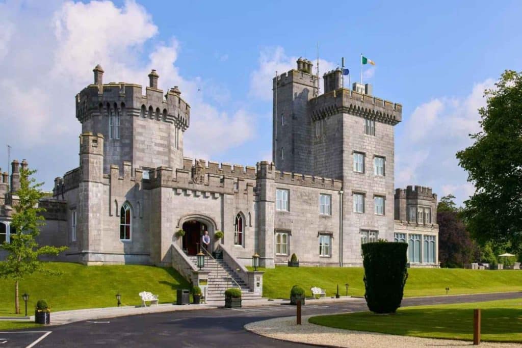 Gray stone castle-style mansion with crenellated towers, arched entrance, and a manicured lawn under a blue sky