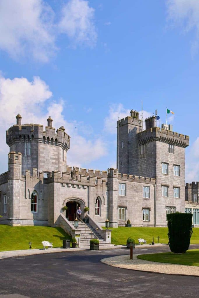 Castle-like stone mansion with two round-topped towers and a grand arched entry on a manicured lawn under a blue sky