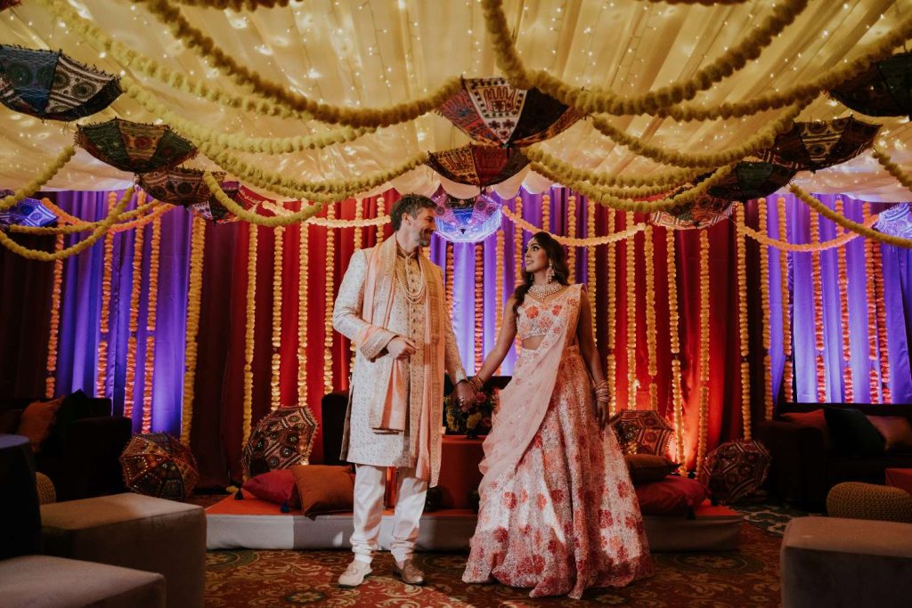 Bride and groom in traditional Indian wedding attire holding hands under a canopy of marigold garlands and fairy lights nearby.