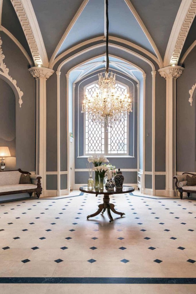 Destination wedding in Ireland at Luttrellstown Castle, County Dublin — the Gothic Hall featuring a black and white tiled floor with a circular table set at the centre, table decorations arranged beneath a chandelier, with natural light streaming through a window behind.