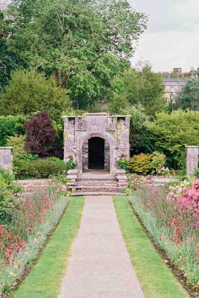 Destination wedding in Ireland at Dromoland Castle, County Clare — stone pathway with steps leading through the walled garden towards an arched opening, bordered by flowers and lawn with mature trees beyond.