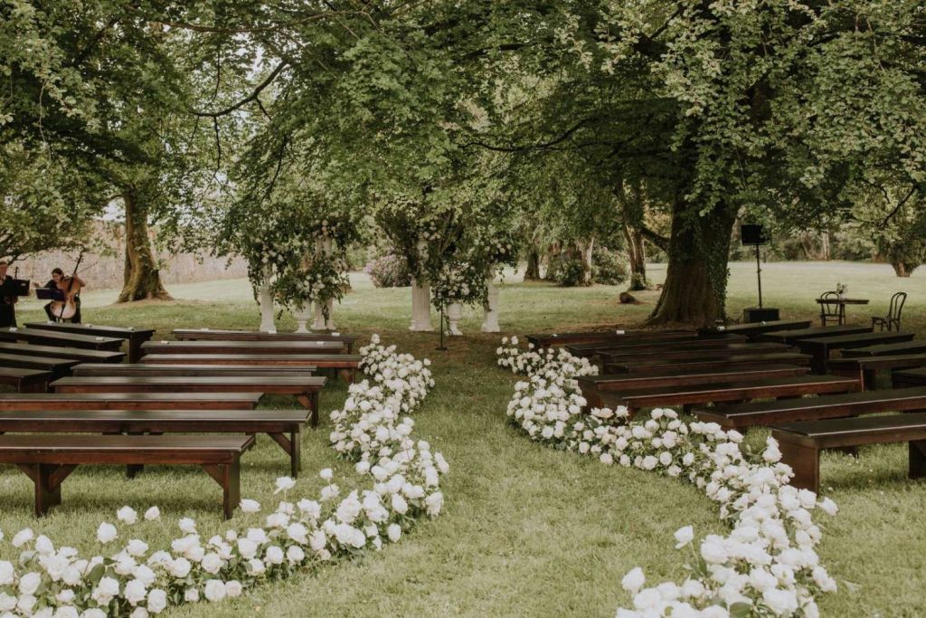 Outdoor garden wedding setup with a white flower-lined aisle and wooden benches under large trees; musicians playing string instruments to the left.
