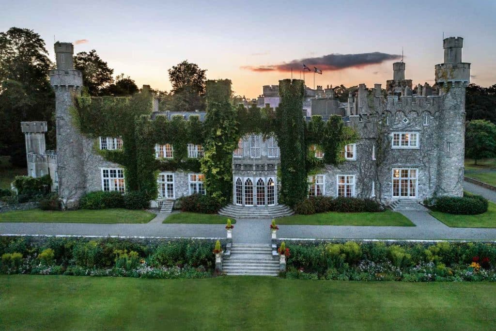 Ivy-covered stone castle with crenellated towers and central bay window, set in formal gardens at sunset.