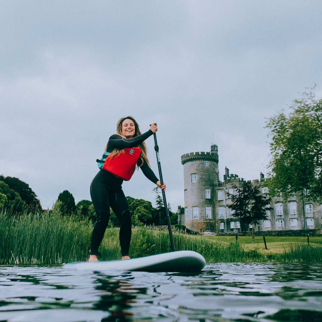 Woman in a red life jacket stand-up paddling on a lake with a castle-like building in the background.