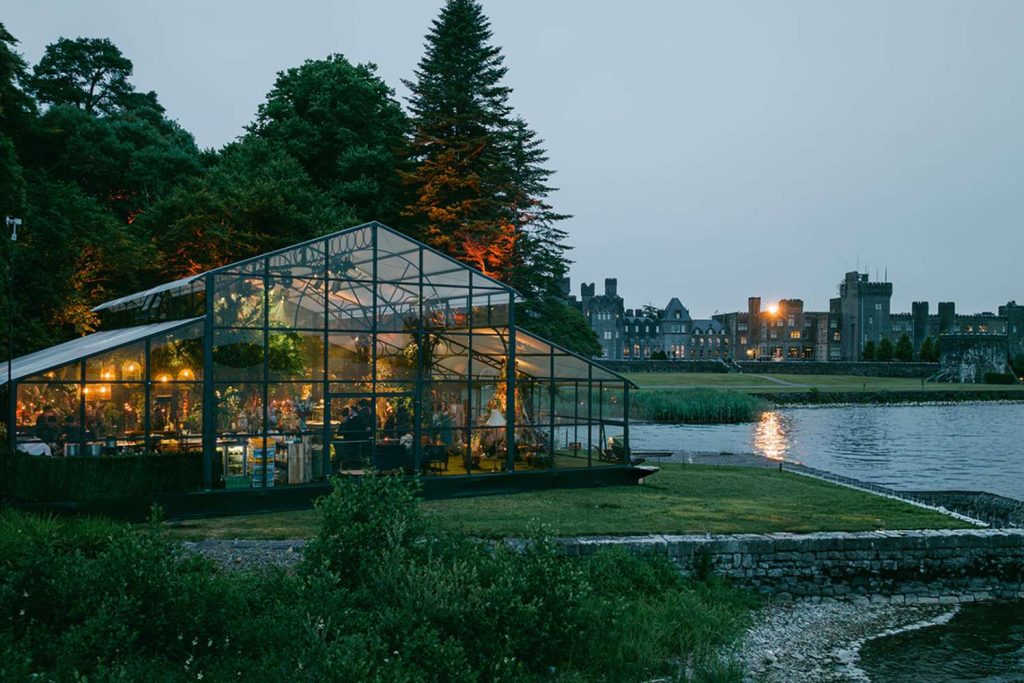 Glass greenhouse restaurant at dusk by a lake, with warm interior lights and a historic castle-like building in the background.