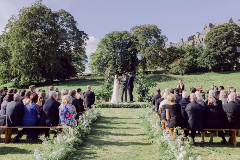 Outdoor wedding ceremony on a sunny lawn with a floral arch as the bride and groom exchange vows, guests seated on benches with a castle in the distance.