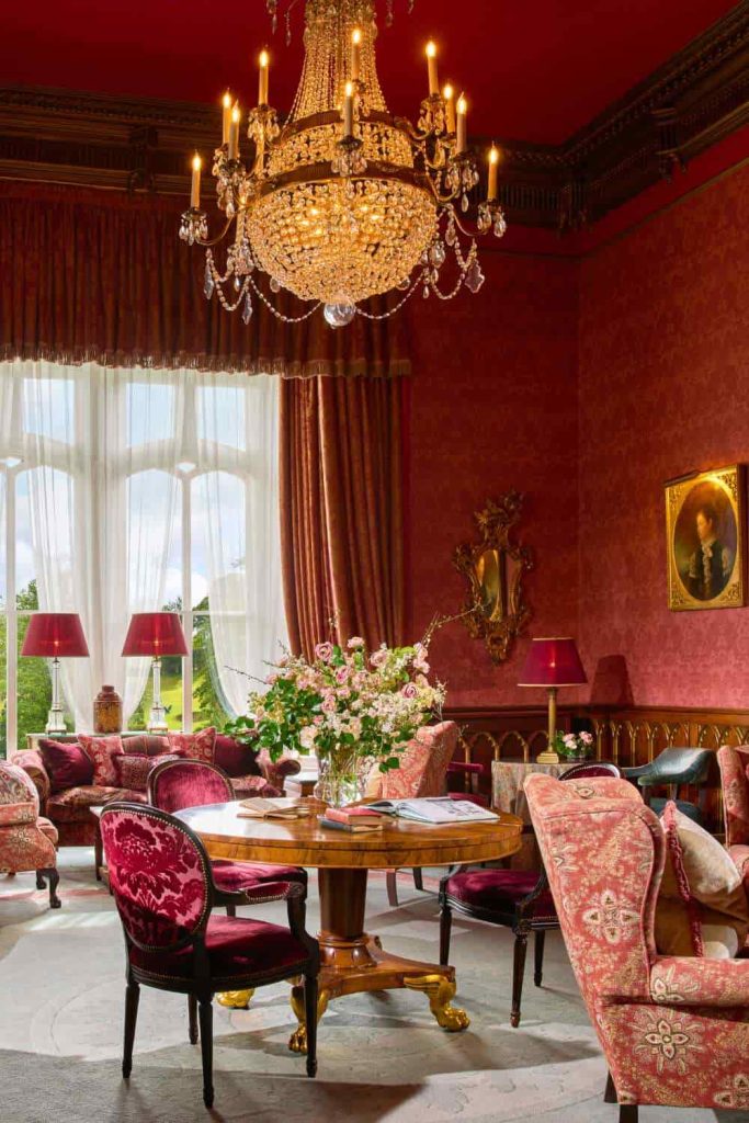 Irish destination wedding at Dromoland Castle, County Clare — The Drawing Room with red wallpaper, chandelier, lounge seating, a circular table with floral arrangement, and upholstered chairs.