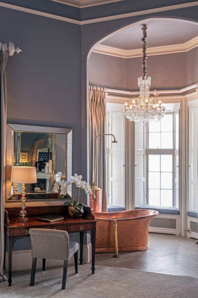 Wedding venue suite at Luttrellstown Castle, County Dublin — The Aileen Plunkett Suite featuring a freestanding copper bathtub beneath a chandelier, with a writing desk, wall mirror, and natural daylight from a nearby window.