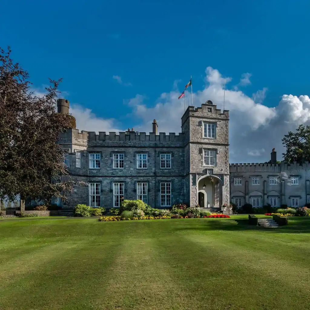 Irish castle estate at Luttrellstown Castle, County Dublin — stone exterior with flag-topped rooflines, arched entrance, and landscaped gardens with flowers and greenery across the front lawn.