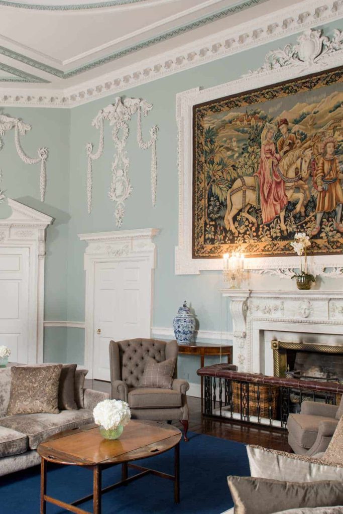 Luttrellstown Castle, County Dublin — The Kentian Room with a high-ceiling interior, fireplace beneath a framed painting, ornate wall detailing, and a lounge seating area with an oval coffee table and floral arrangement.
