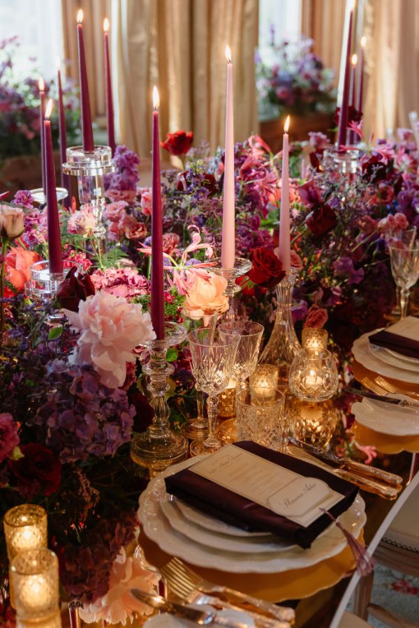Elegant dining table with tall pink and burgundy candles, crystal glassware, and a lush floral centerpiece.
