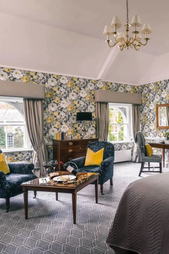 Irish destination wedding at Mount Juliet Estate, County Kilkenny, Ireland — Garden View King Room featuring upholstered armchairs, a table with cheese board and wine, floral patterned wallpaper, and draped window curtains.