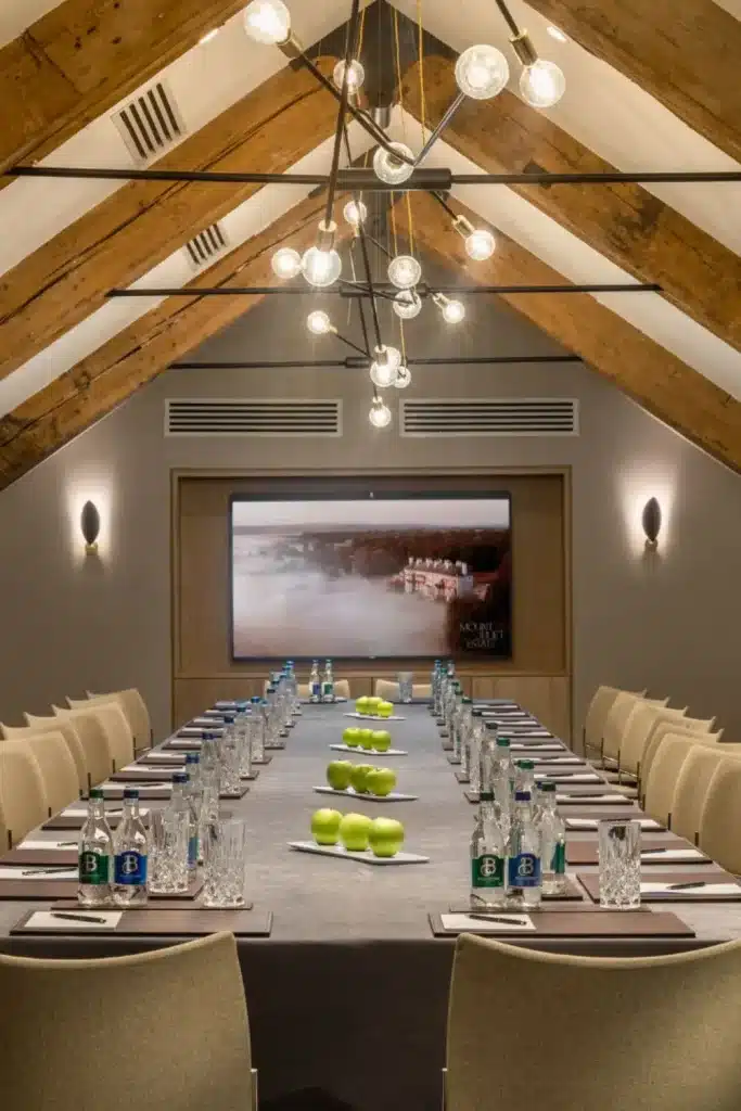 Destination wedding at Mount Juliet Estate, County Kilkenny — Jack Nicklaus Boardroom set with a long meeting table, place settings including notepads, pens, water glasses, and fresh green apples arranged along the centre, with a wall-mounted screen at the end of the room.