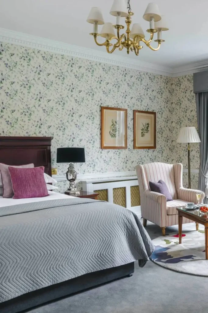Destination wedding accommodation at Mount Juliet Estate, County Kilkenny, Ireland — Junior Suite with a plush bed and layered pillows, floral patterned wallpaper with framed artwork, upholstered armchair, side table, and standing lamp.