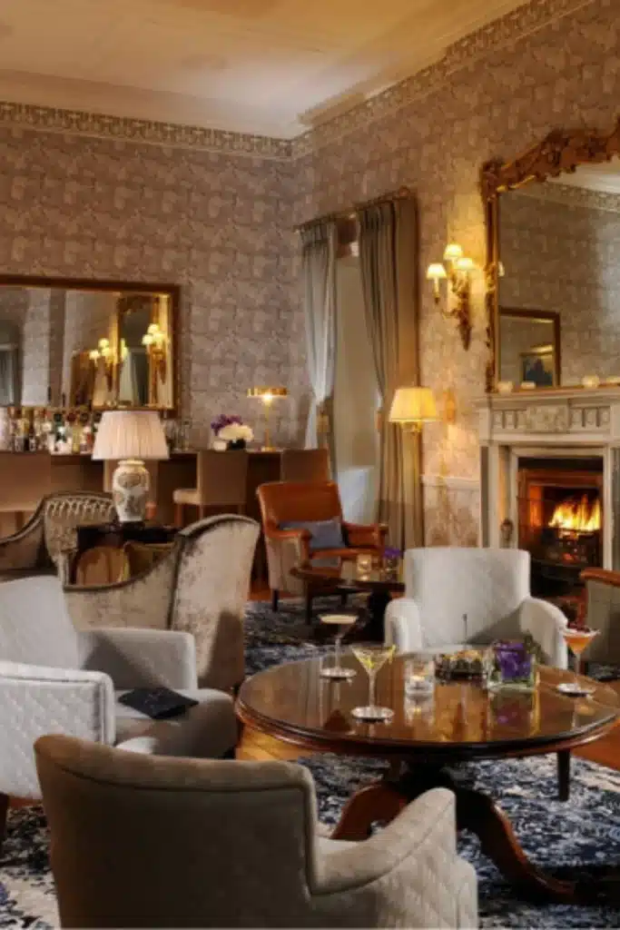 Irish destination wedding at Mount Juliet Estate, County Kilkenny, Ireland — The Major’s Bar lounge with armchairs around a round table, cocktails on the table, soft lamplight, a lit fireplace, and patterned wallpaper with a mirror above the mantel.