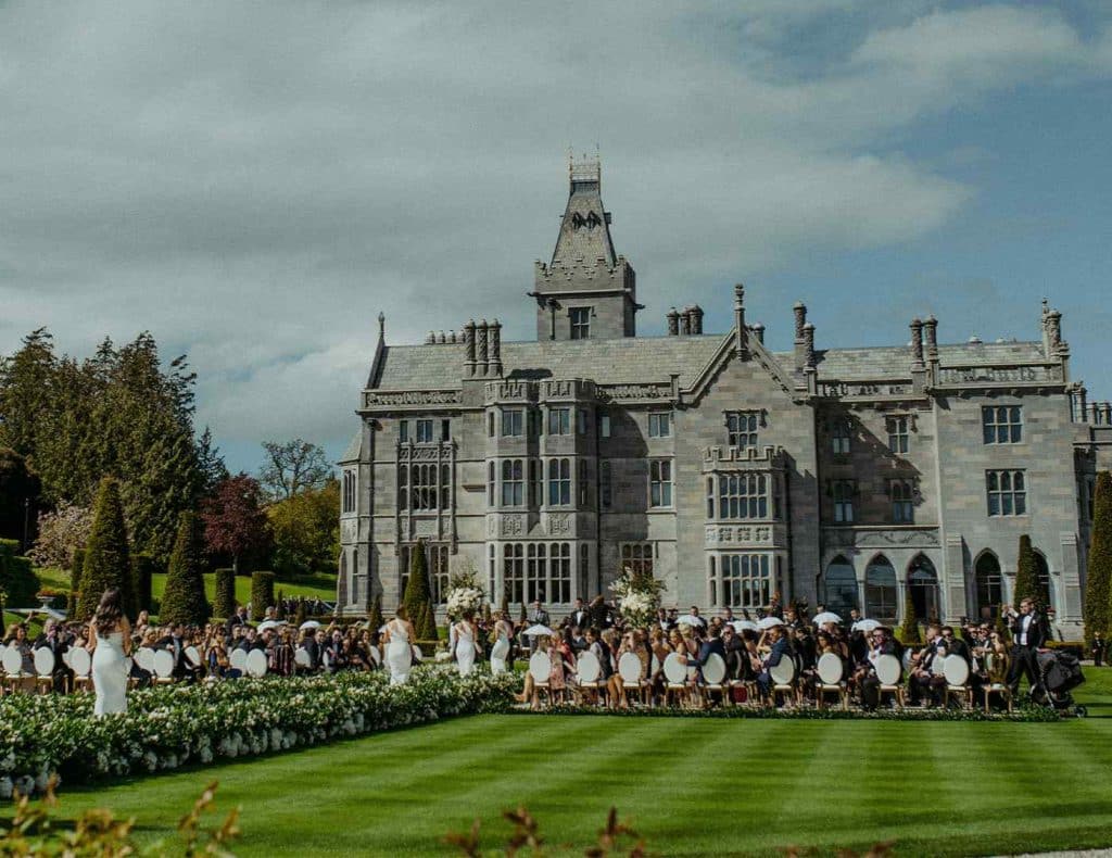 Wedding ceremony on a manicured lawn in front of a large historic mansion, guests seated and bridesmaids in white dresses.